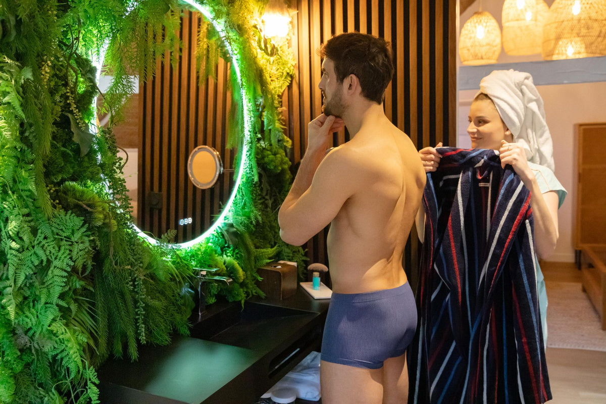 Un homme en sous-vêtement bleu se tient devant un miroir de salle de bain tandis qu'une femme en serviette lui enroule un peignoir rayé