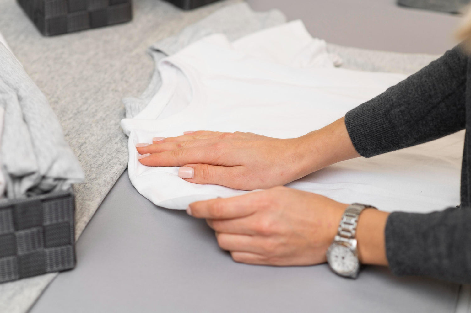 Plan rapproché d'une personne pliant soigneusement un t-shirt blanc, avec des piles de vêtements gris en arrière-plan.